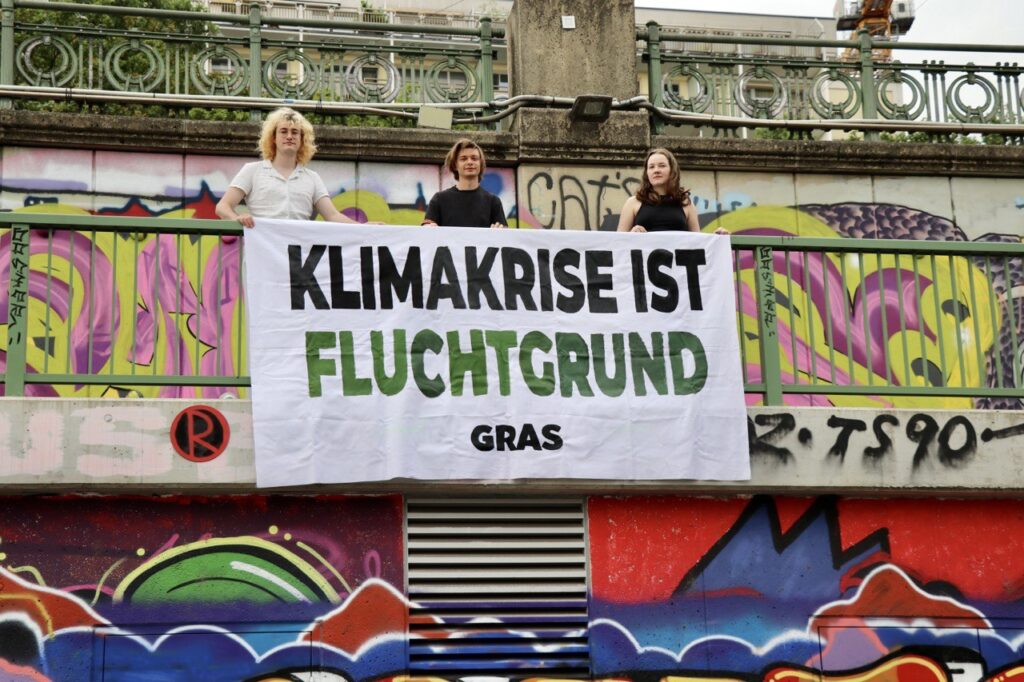 Drei Demonstrierende mit einem Banner mit der Aufschrift "Klimakrise ist Fluchtgrund" vor einer Wand mit Graffitis.